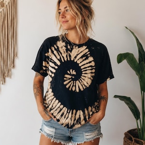 May include: A black short-sleeve t-shirt featuring a large, off-center beige spiral tie-dye pattern across the front. The shirt has a crew neckline and short sleeves with rolled cuffs, paired with light blue denim shorts with frayed hems.