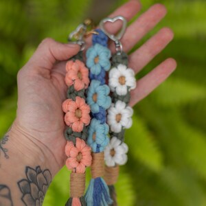 May include: Handmade macrame keychains with flower accents. Each keychain features a series of colorful, woven flowers in shades of peach, blue, and white. The keychains have a tassel and a metal heart-shaped clasp.