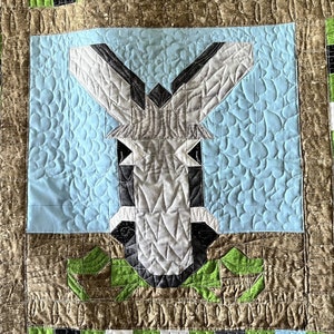 May include: A quilt with a patchwork design featuring a gray donkey in the center. The quilt is made with a variety of fabrics in shades of green, gray, black, and white. The donkey is surrounded by a blue background with a quilted pattern.