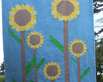 Könnte beinhalten: Eine blaue Quiltdecke mit fünf aufgenähten Sonnenblumen. Die Sonnenblumen sind gelb mit braunen Zentren und grünen Blättern. Die Decke hängt an einer Wäscheleine.