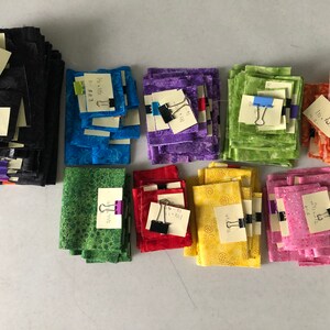 K&ouml;nnte beinhalten: Stapel von Stoffquadraten in verschiedenen Farben, darunter Schwarz, Blau, Lila, Gr&uuml;n, Rot, Gelb und Pink. Jeder Stapel hat einen handgeschriebenen Zettel mit einer B&uuml;roklammer befestigt.
