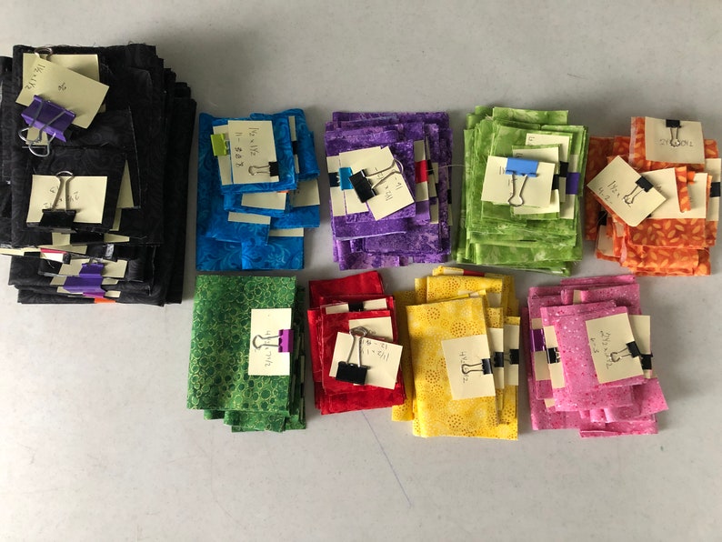 K&ouml;nnte beinhalten: Stapel von Stoffquadraten in verschiedenen Farben, darunter Schwarz, Blau, Lila, Gr&uuml;n, Rot, Gelb und Pink. Jeder Stapel hat einen handgeschriebenen Zettel mit einer B&uuml;roklammer befestigt.