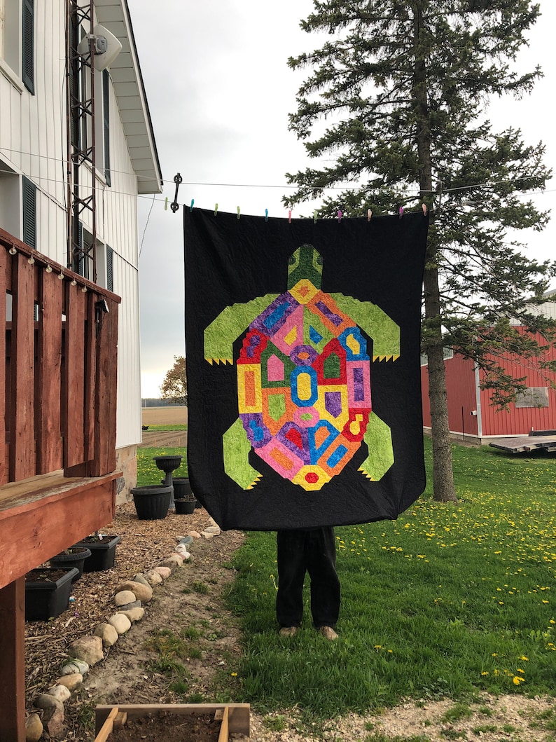 K&ouml;nnte beinhalten: Eine schwarze Steppdecke mit einem bunten geometrischen Schildkr&ouml;tenmuster. Die Steppdecke h&auml;ngt an einer W&auml;scheleine vor einem Haus und einem Baum.