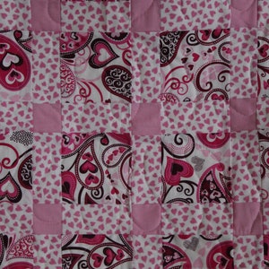 Peut inclure: Une couverture matelassée avec un motif répétitif de cœurs roses et de motifs cachemire sur fond blanc. La couverture est bordée d'un tissu rose uni et présente un motif cousu.