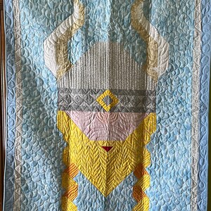 May include: A blue quilted wall hanging with a yellow, pink, and gray Viking helmet design. The helmet has a diamond shape in the center and braids on the sides.