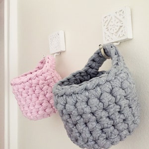 May include: Two crocheted hanging baskets, one pink and one gray, are hung on white decorative hooks on a white wall.