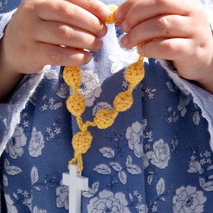 May include: A child's hands holding a yellow crocheted rosary with a white cross at the end.