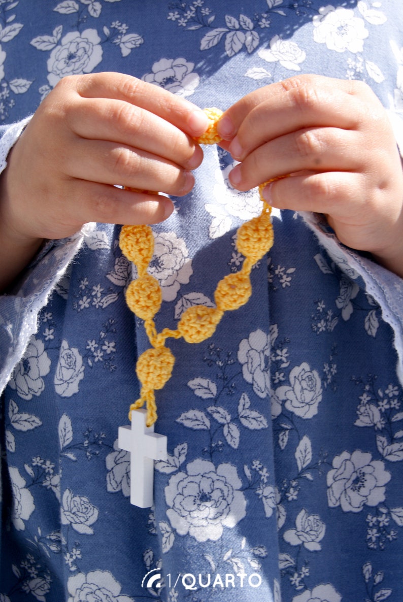 May include: A child's hands holding a yellow crocheted rosary with a white cross at the end.