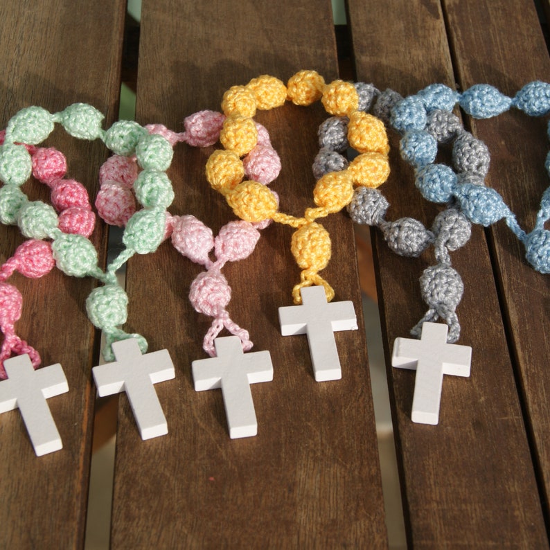 May include: Five crocheted rosaries with wooden crosses. The rosaries are in different colors: pink, green, yellow, blue, and gray.
