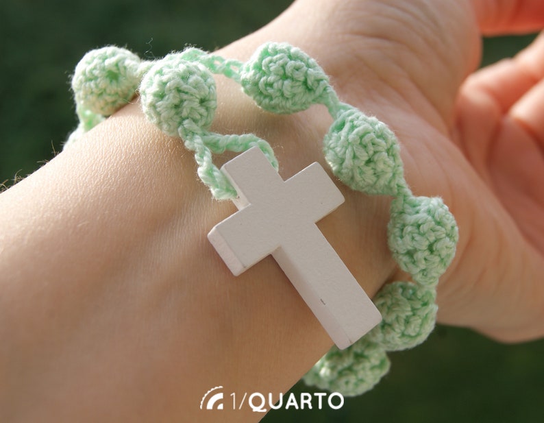 May include: A green crocheted bracelet with a white wooden cross charm. The bracelet is made with small, round beads and has a simple design.