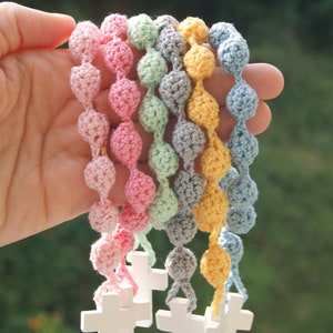 May include: A set of seven crocheted beaded necklaces with wooden cross charms. The necklaces are in pastel colors: pink, light blue, green, gray, and yellow.