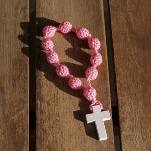 May include: A pink crocheted rosary with a white wooden cross. The rosary is laid out on a wooden surface.