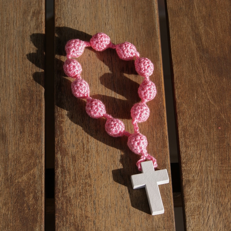 May include: A pink crocheted rosary with a white wooden cross. The rosary is laid out on a wooden surface.