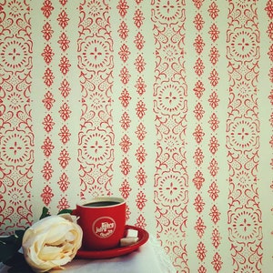 Pode incluir: Um fundo branco com um papel de parede estampado em vermelho e branco. Um pires de cerâmica branco com uma xícara de café vermelha em cima. A xícara de café tem as palavras "Dobro Jutro" impressas. Uma rosa branca é parcialmente visível no canto inferior esquerdo da imagem.