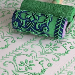 May include: A green patterned paint roller with a floral design sits atop a white surface with a matching green floral pattern. The roller has a blue and green design. The surface has a repeating floral and vine pattern.