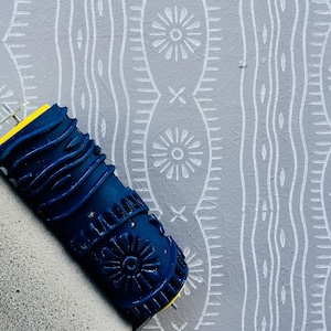 May include: A blue and yellow paint roller with a foam handle is being used to create a white patterned design on a gray wall.