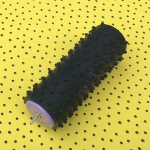 May include: A black massage roller with a textured surface and small, rounded protrusions. The roller has a light purple end cap and a metal rod extending from it. The roller is set against a yellow background with black polka dots.