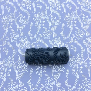 May include: A black paint roller with a floral design. The roller is on a purple background with a white floral pattern.