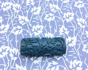 May include: A dark blue paint roller with a floral design. The roller is on a light purple background with a white floral pattern.