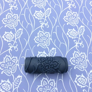May include: A black patterned paint roller with a floral design is displayed against a lavender background with a matching floral pattern. The roller is designed for creating decorative wall finishes. The floral pattern includes paisley and leaf elements.