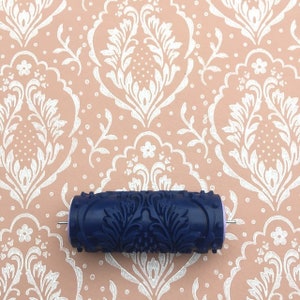 Puede incluir: Un rodillo de pintura con diseño floral azul oscuro, sobre un fondo de patrón de damasco rosa y blanco. El rodillo está diseñado para crear patrones decorativos en paredes. La imagen muestra una herramienta para la decoración del hogar y proyectos de bricolaje.