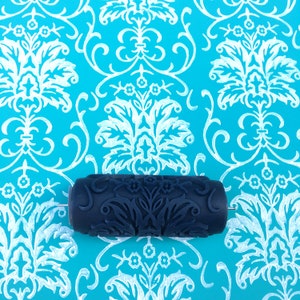 May include: A dark blue paint roller with a decorative pattern, designed to create a damask pattern on a wall. The roller is on a blue background with a white damask pattern.