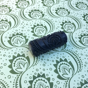 May include: A dark brown patterned paint roller with a floral design, resting on a light green and white floral patterned surface. The roller has a metal pin extending from one end. The background features a repeating floral pattern.
