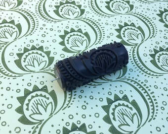 May include: A dark brown patterned paint roller with a floral design, resting on a light green and white floral patterned surface. The roller has a metal pin extending from one end. The background features a repeating floral pattern.