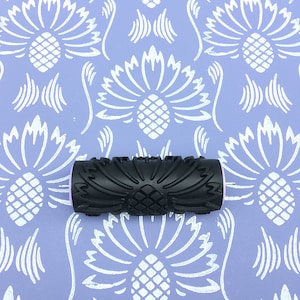 May include: A black rubber paint roller with a detailed floral design. The roller is on a purple background with a white floral pattern.