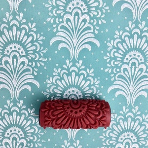 May include: A red patterned paint roller rests on a teal surface with a white floral design. The roller features a repeating, symmetrical pattern. The background has a repeating white floral design on a teal background.