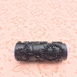May include: A black paint roller with a floral design. The roller is made of plastic and has a metal handle. The roller is on a pink background with a floral pattern.