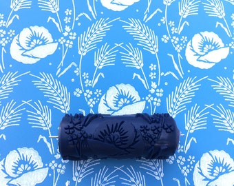 Op de afbeelding: Een blauwe verfroller met een bloemenpatroon. De roller ligt op een blauwe achtergrond met een wit bloemenpatroon.
