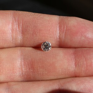 Puede incluir: Una sola piedra preciosa de diamante redonda y transparente sostenida en una mano.