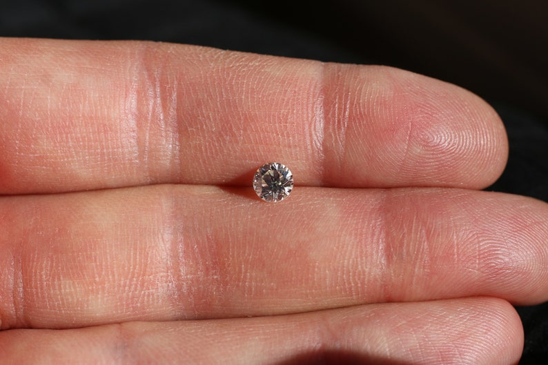 Puede incluir: Una sola piedra preciosa de diamante redonda y transparente sostenida en una mano.