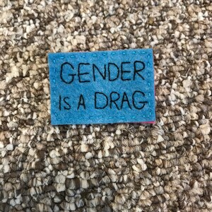 Op de afbeelding: Een blauwe viltpatch met witte stiksels rond de randen. De tekst "GENDER IS A DRAG" is geborduurd in zwart garen.