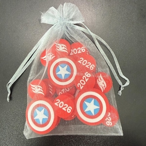 May include: A collection of red and white circular tokens with a blue star design and the number "2026" printed on them. The tokens are contained within a sheer, light blue drawstring bag.