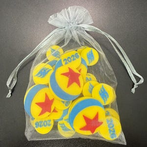 May include: A collection of yellow, blue, and red circular objects with a star and flame design, and the number 2026, inside a sheer, light blue drawstring bag. The objects are scattered within the bag.