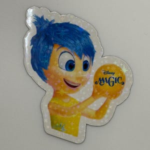 May include: A holographic sticker featuring the character Joy from the Disney Pixar movie Inside Out. Joy has blue hair and is holding a yellow orb with the word "MAGIC" on it. The sticker has a star-shaped border.