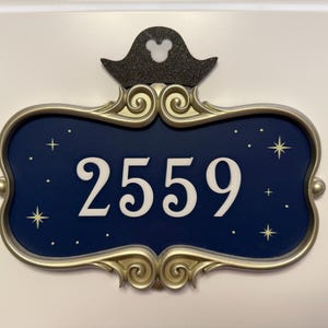 May include: Decorative room number sign with a navy blue background and the number 2559 in white. The sign has a gold-colored ornate frame and is topped with a black pirate hat with a Mickey Mouse silhouette.
