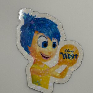 May include: A holographic sticker featuring the character Joy from Disney's "Inside Out" holding a golden orb with the "Disney Wish" logo. Joy has blue hair and a yellow dress, with a smiling expression.