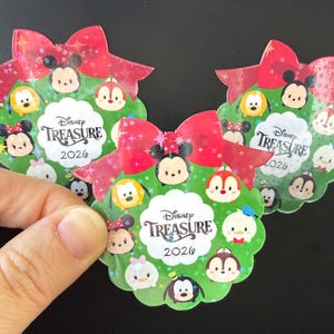May include: Three Disney Treasure 2026 stickers. Each sticker features a green wreath design with various Disney Tsum Tsum characters, a red bow, and the text "Disney Treasure 2026". One sticker is being held by a hand.