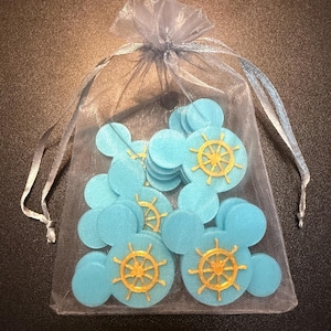 May include: A collection of light blue, Mickey Mouse-shaped confetti with gold ship wheel designs, presented in a sheer, gray drawstring bag. The confetti is scattered within the bag, creating a playful and thematic nautical design.
