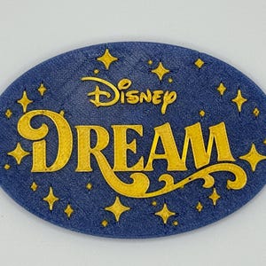 May include: An oval-shaped, navy blue emblem with the word "DREAM" in gold, stylized lettering. The Disney logo is above the word "DREAM". Gold stars are scattered around the edge of the emblem. The emblem has a textured surface.