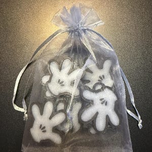 May include: A collection of white cartoon hand-shaped charms with black outlines and details, presented in a sheer, light blue drawstring bag. The bag is tied with a matching ribbon, creating a cohesive and whimsical presentation.