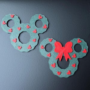 May include: Two green Mickey Mouse-shaped wreaths decorated with small red hearts. One wreath has a large red bow. The wreaths are made of a textured material and are displayed on a dark surface.
