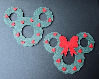 May include: Two green Mickey Mouse-shaped wreaths decorated with small red hearts. One wreath has a large red bow. The wreaths are made of a textured material and are displayed on a dark surface.