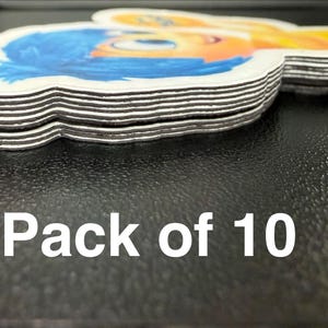 May include: A stack of ten character-shaped magnets featuring a cartoon character with blue hair and a smiling face. The magnets have a white border and are stacked on a dark surface. The text "Pack of 10" is visible.