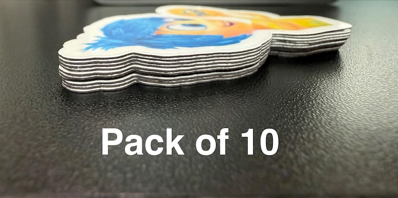 May include: A stack of ten character-shaped magnets featuring a cartoon character with blue hair and a smiling face. The magnets have a white border and are stacked on a dark surface. The text "Pack of 10" is visible.