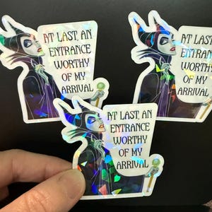 May include: Three holographic stickers featuring a stylized illustration of a character with dark horns and a high-collared robe, holding a staff. The text reads: AT LAST, AN ENTRANCE WORTHY OF MY ARRIVAL. The stickers have a reflective finish.