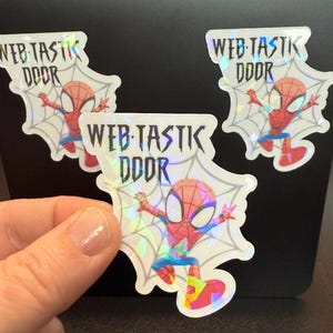 May include: Holographic stickers featuring Spider-Man in a web design. The stickers have the text "WEB-TASTIC DOOR" in a stylized font. The Spider-Man character is in a classic pose, with red and blue colors.